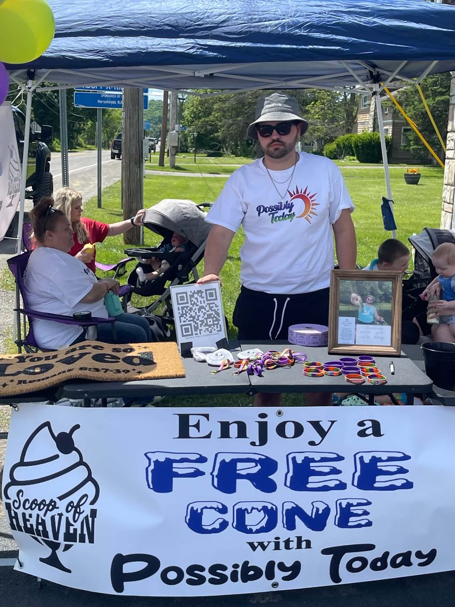 The Scoop of Heaven tent with a banner reading Enjoy a free cone with Possibly Today, and a table of wristbands, a doormat and a QR code for the Facebook group
